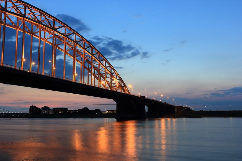 De Waalbrug bij Nijmegen, verlicht in de avondschemering, met de rivier op de voorgrond